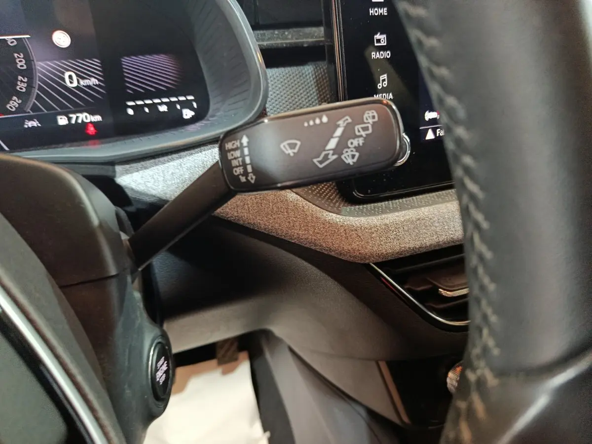 Gros plan sur la commande d’essuie-glace à gauche du volant cuir, avec tableau de bord digital et écran tactile en arrière-plan.