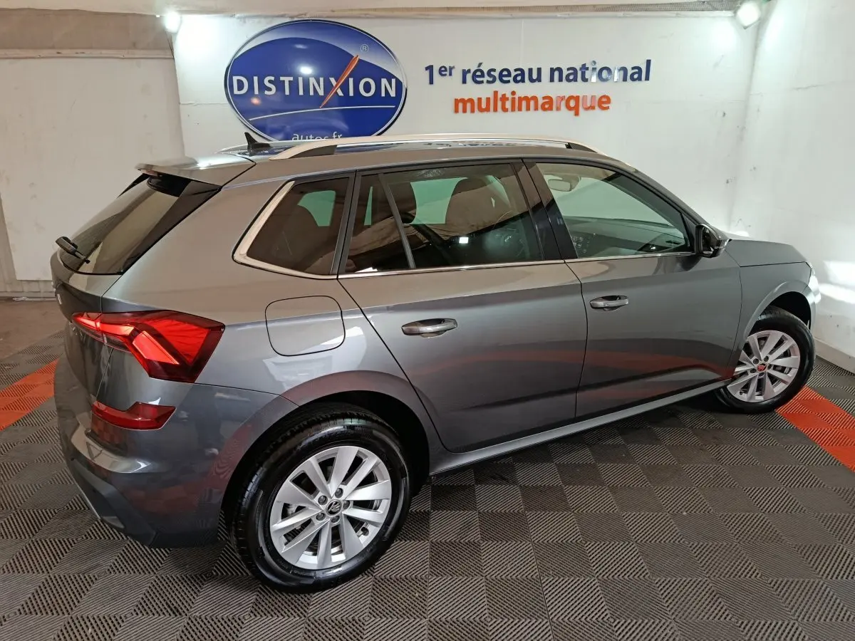 Vue 3/4 arrière droite d’un Skoda Kamiq gris clair 2024 avec jantes alliage et barres de toit noires.