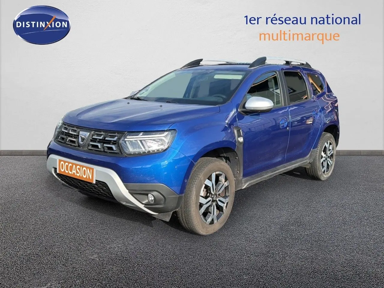 Dacia Duster bleu Iron en 3/4 avant droit avec jantes bi-ton et barres de toit visibles.