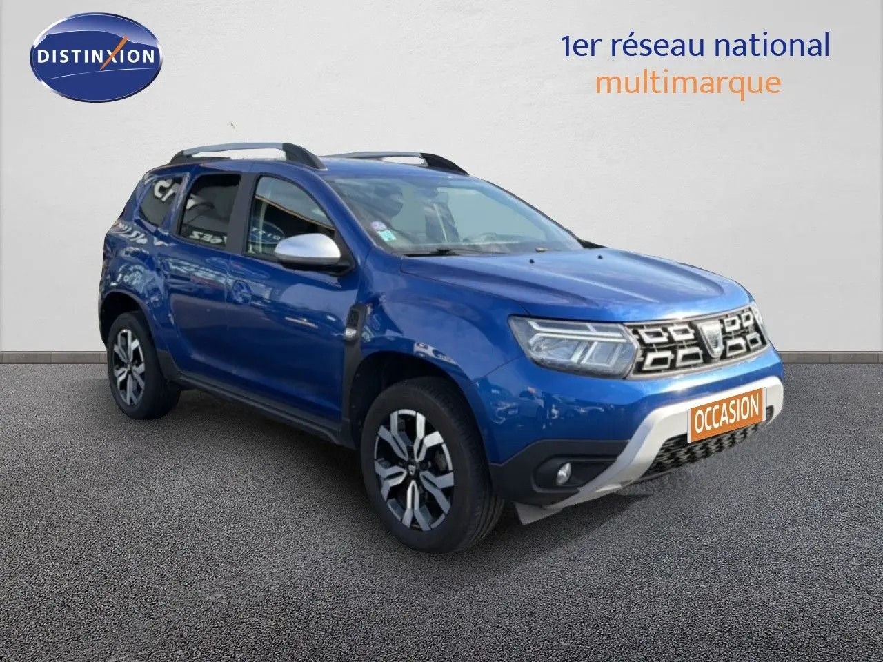 Dacia Duster bleu Iron en 3/4 avant droit, SUV compact avec jantes bi-ton et barres de toit visibles.