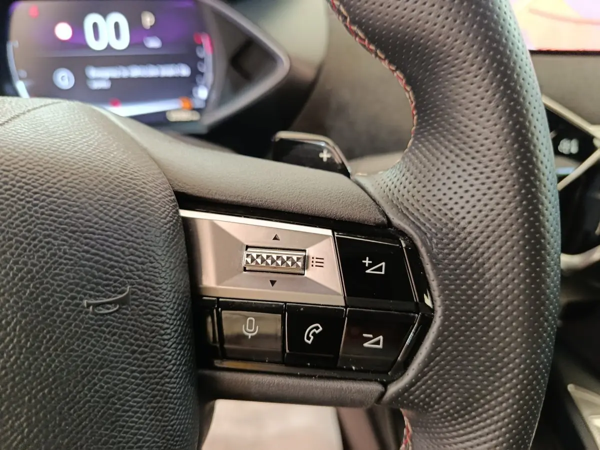Gros plan sur le volant cuir perforé du DS3 2024 avec commandes vocales et téléphone, combiné numérique flou en arrière-plan.