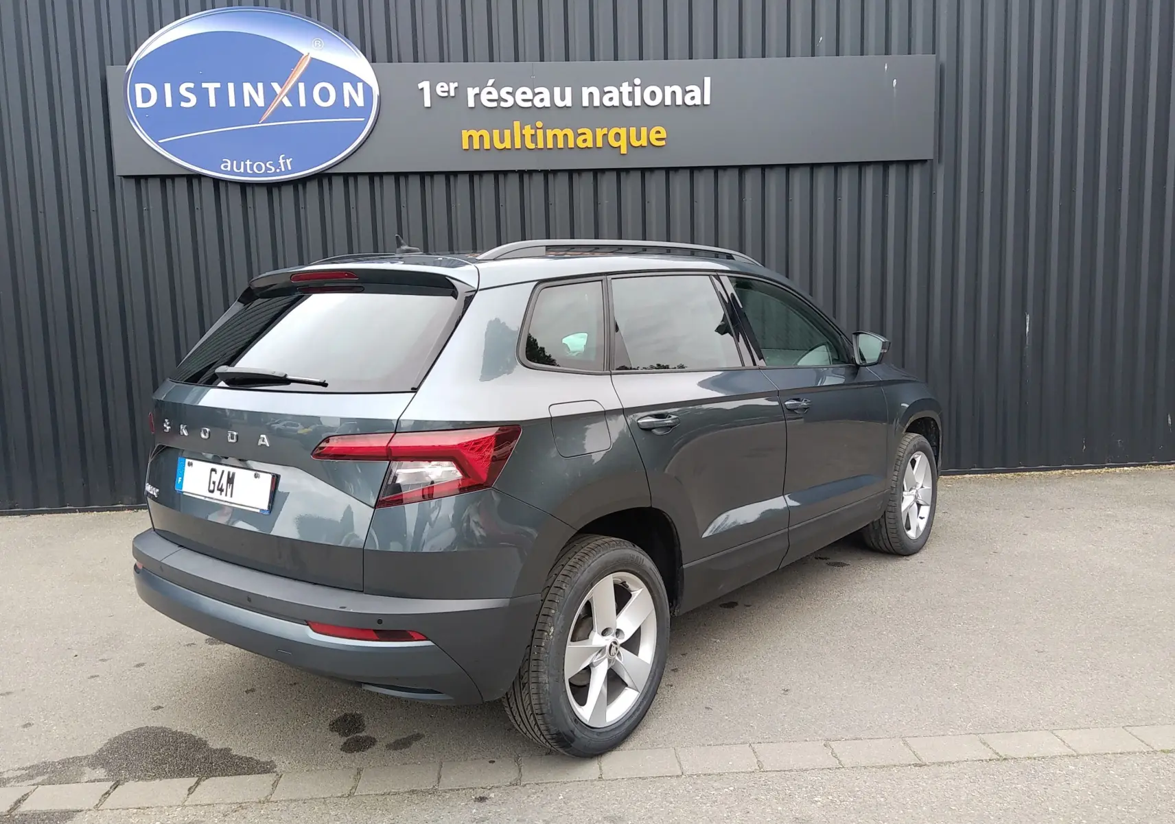 Vue 3/4 arrière droite d'un Skoda Karoq gris 2021 garé devant un mur noir avec un panneau Distinxion.