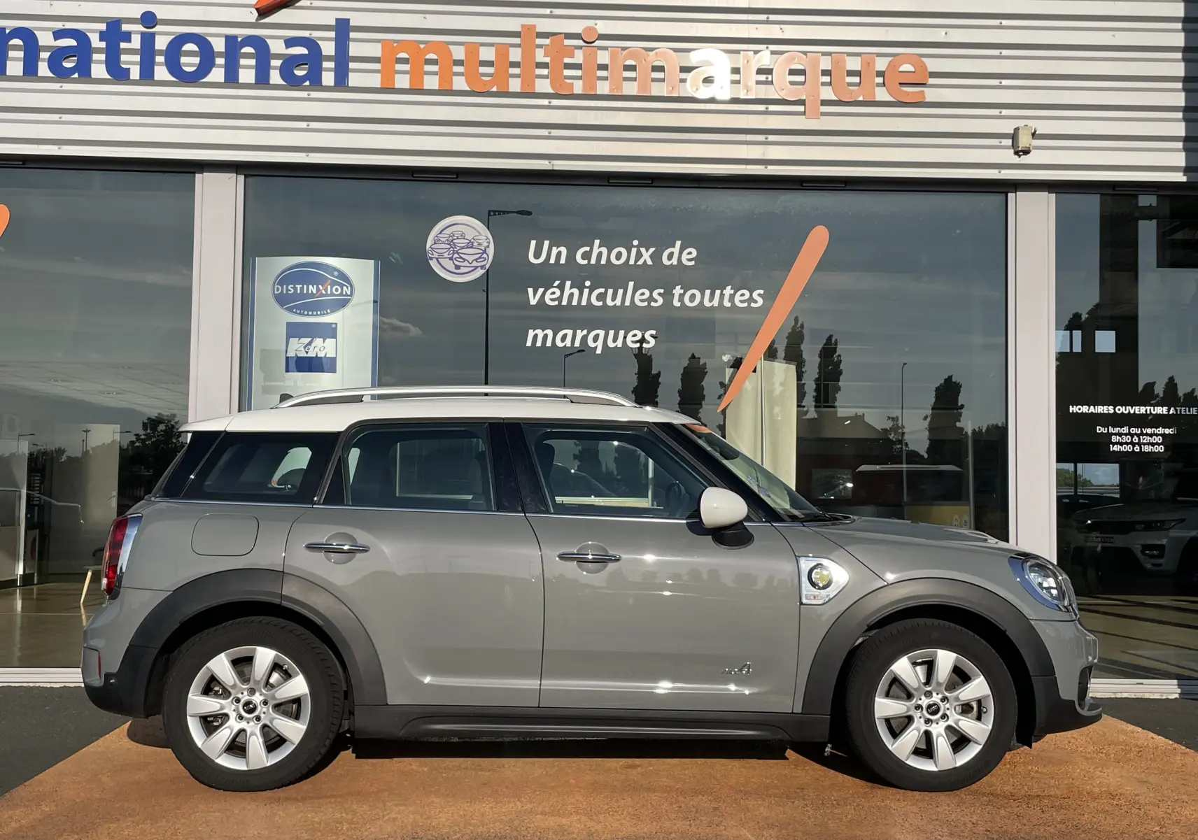 Profil côté droit d'une MINI COUNTRYMAN F60 hybride rechargeable Moonwalk Grey garée devant une vitrine multimarque.
