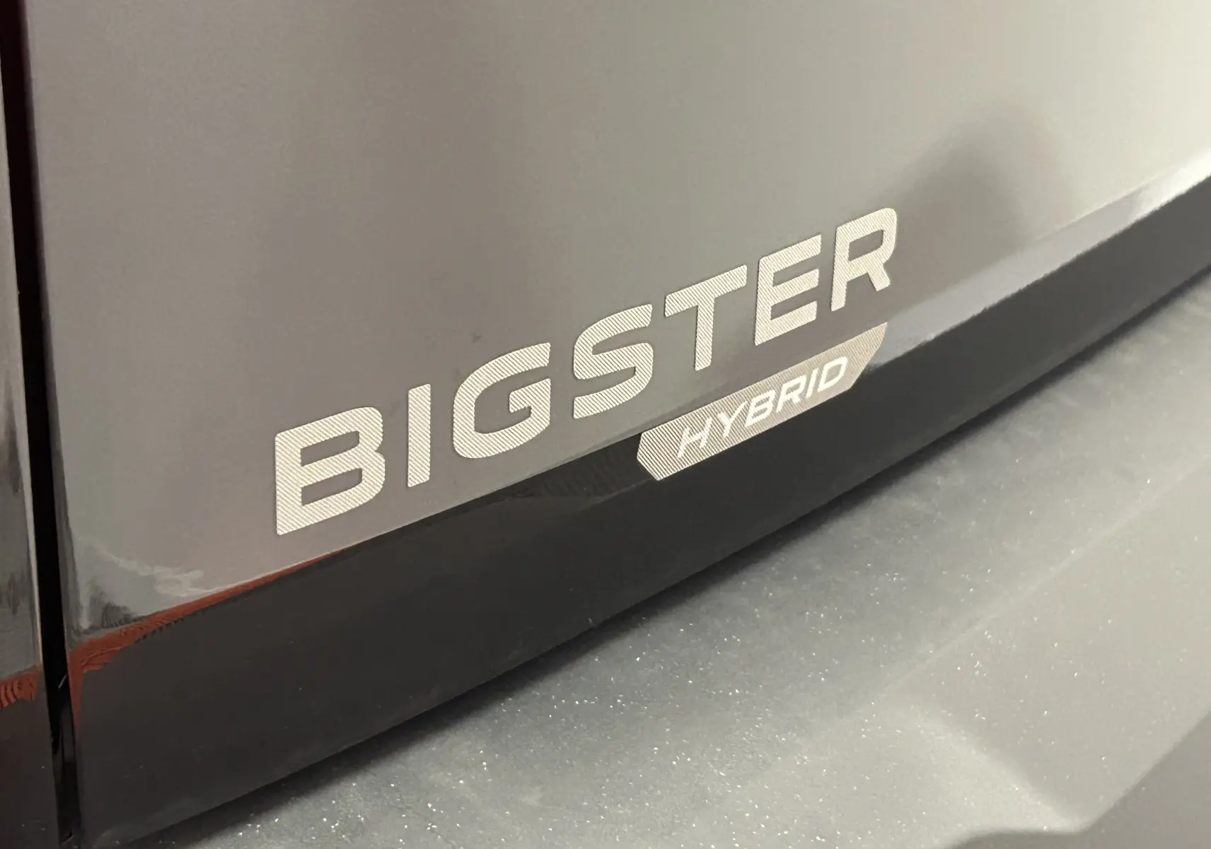 Gros plan sur l’inscription « BIGSTER HYBRID » en blanc sur la carrosserie noir nacré du véhicule.