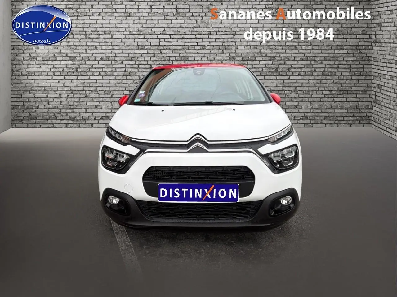 Vue frontale d'une Citroën C3 blanche avec toit et rétroviseurs rouges, sur fond de mur en briques grises.