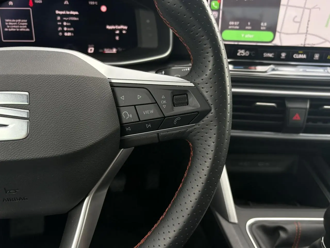 Gros plan sur le volant en cuir noir perforé avec surpiqûres rouges du SEAT Leon 2025, tableau de bord et écran tactile en arrière-plan.