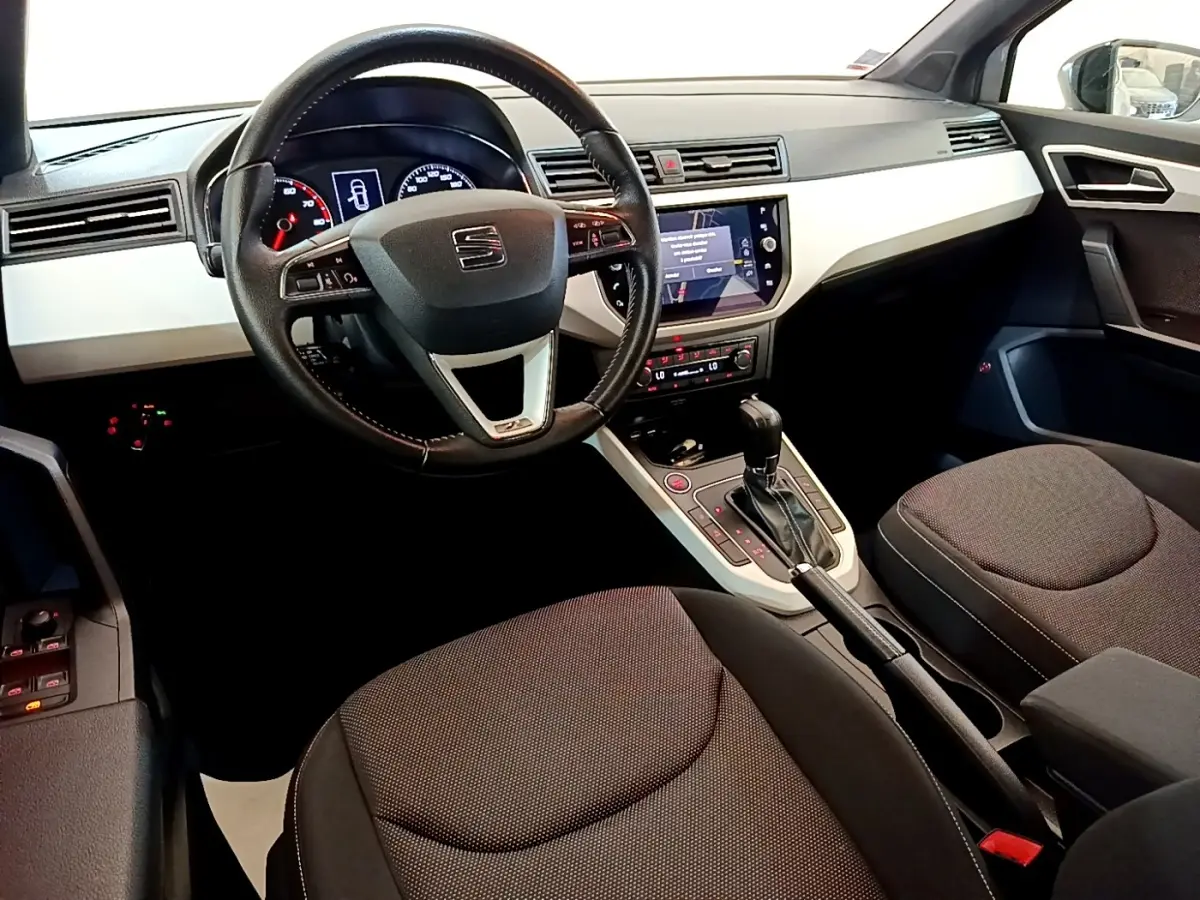 Vue intérieure avant du tableau de bord et volant cuir alu d’un SEAT Arona gris clair avec écran tactile central allumé.