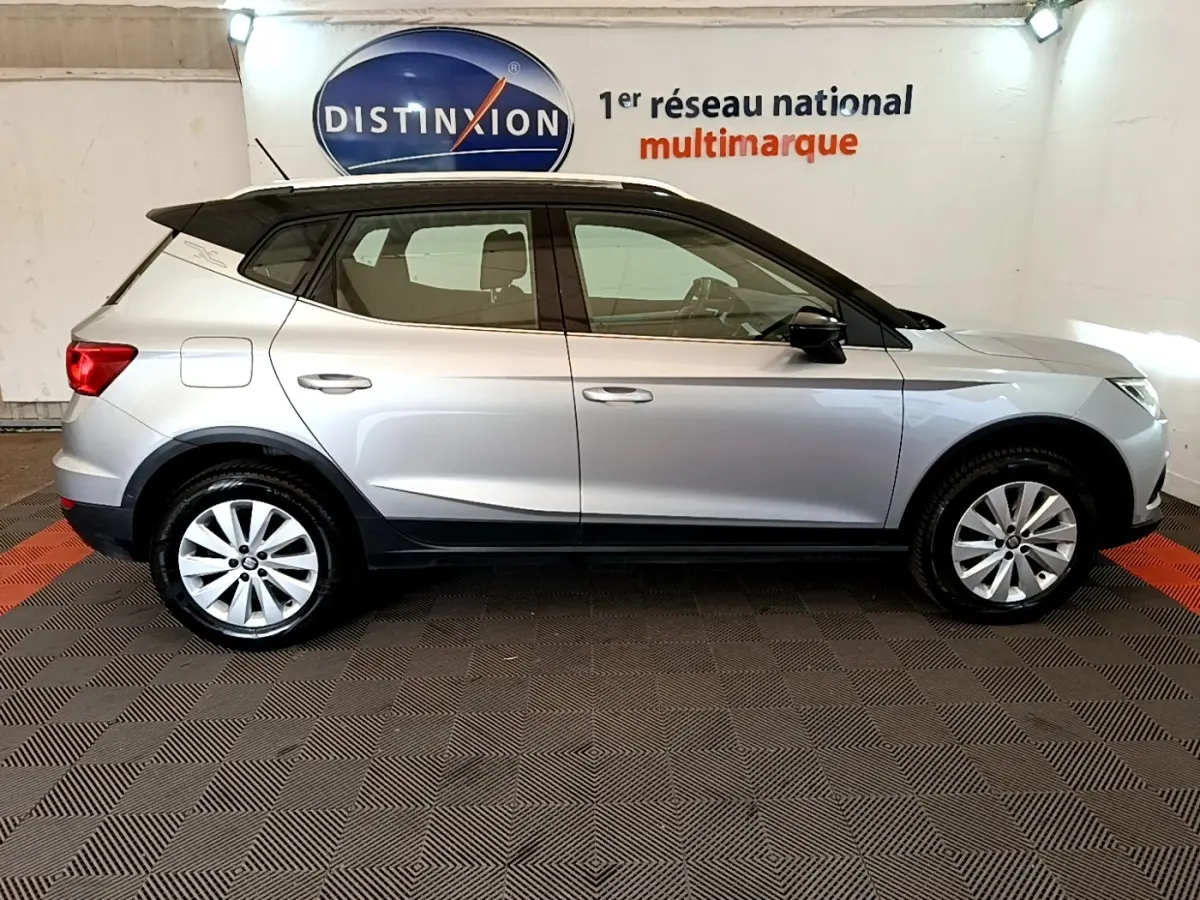Vue de profil côté gauche d'un SEAT Arona gris clair avec toit noir, dans un showroom intérieur.