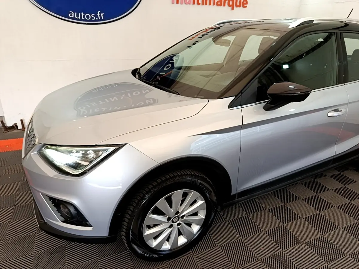 SEAT Arona gris clair vu de 3/4 avant droit, avec phares LED et jantes alliage visibles en intérieur showroom.