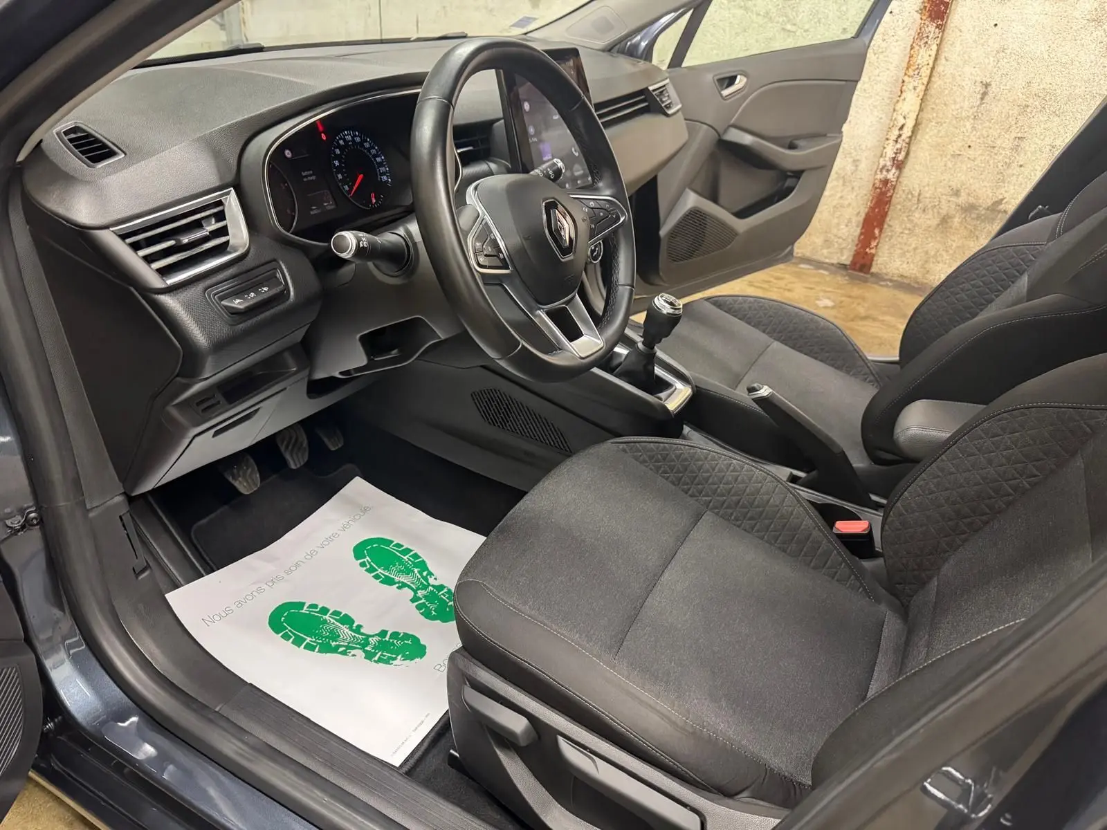 Vue intérieure côté conducteur de la Renault Clio V Business gris titanium, montrant le volant, la boîte manuelle et les sièges en tissu noir.