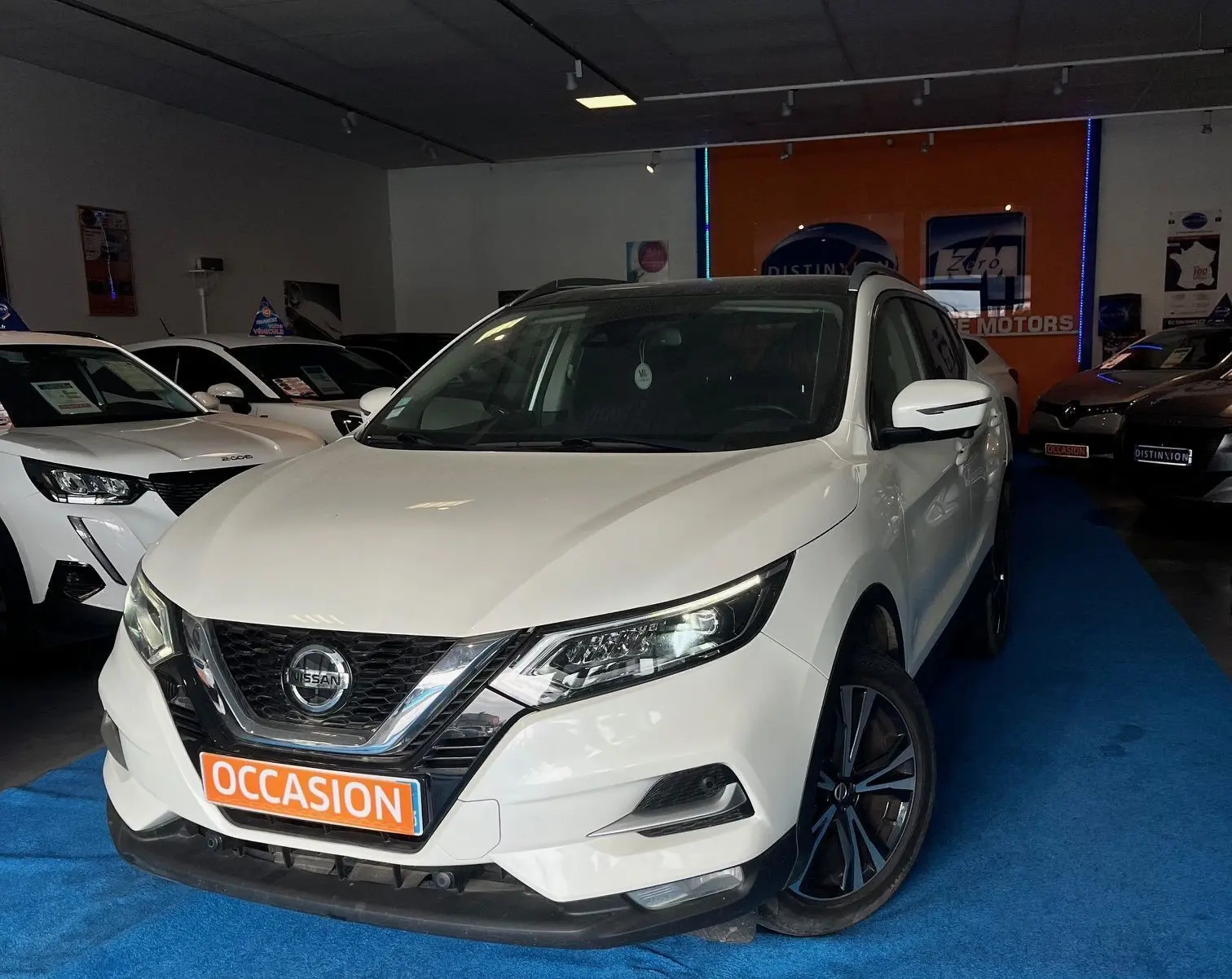 Nissan Qashqai blanc lunaire en 3/4 avant droit, avec jantes noires et plaque "OCCASION" en showroom.