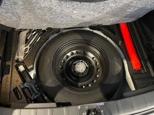 Vue plongeante sur la roue de secours noire du coffre d'un Nissan Qashqai blanc lunaire, avec cric et outils rangés autour.
