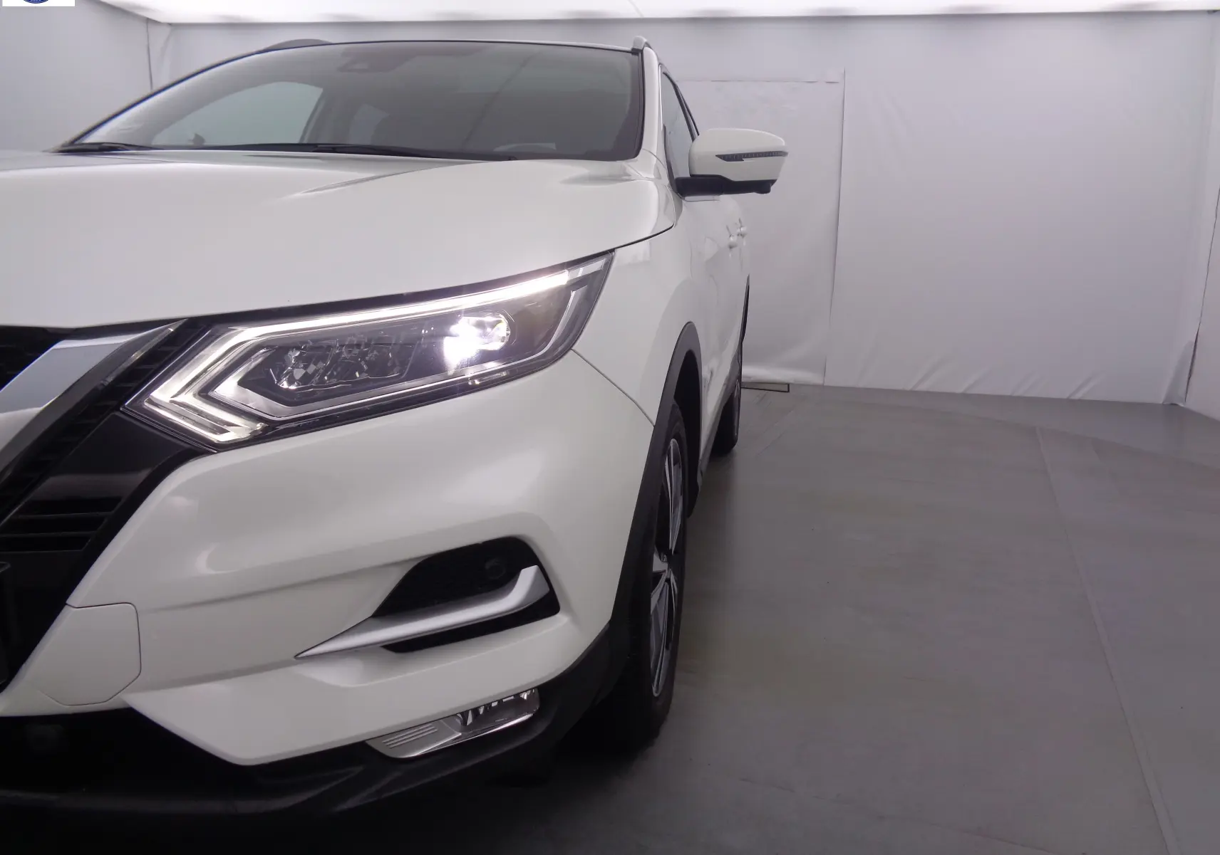 Vue 3/4 avant droit du Nissan Qashqai blanc lunaire métal avec phares allumés dans un studio lumineux.