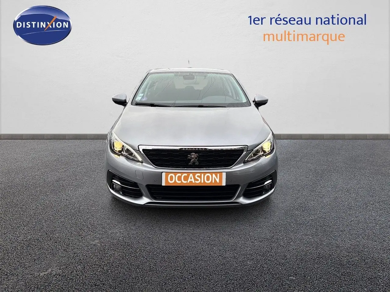 Vue frontale d'une Peugeot 308 gris artense avec phares allumés et plaque d'immatriculation orange "OCCASION" visible.