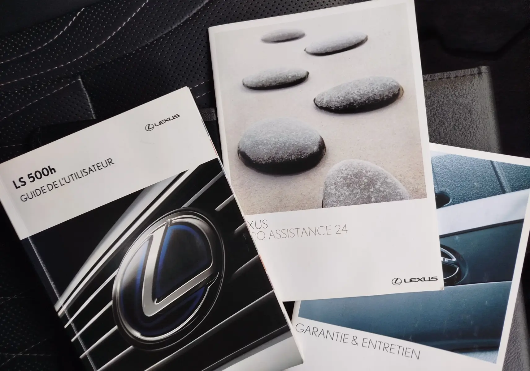 Vue rapprochée de trois brochures Lexus posées sur un siège en cuir noir perforé, sans véhicule visible.