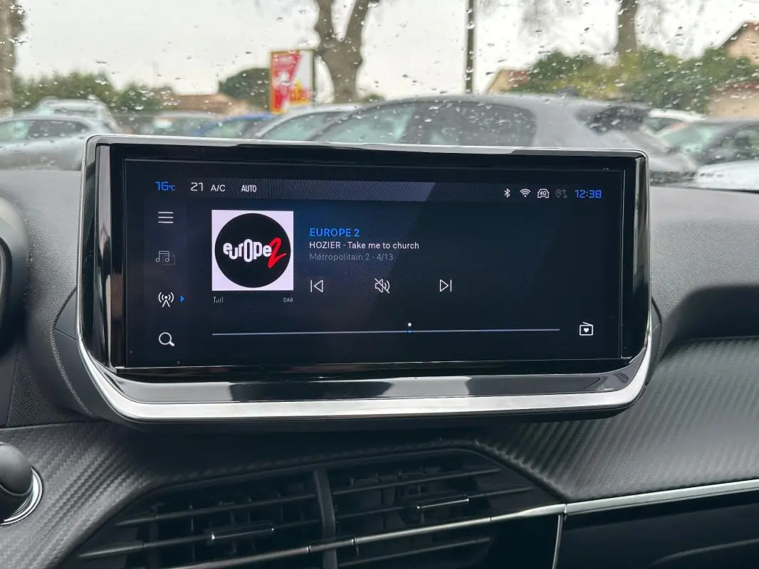 Écran tactile central du tableau de bord du Peugeot 2008 Hybrid 145 Allure, affichant une radio en extérieur pluvieux.