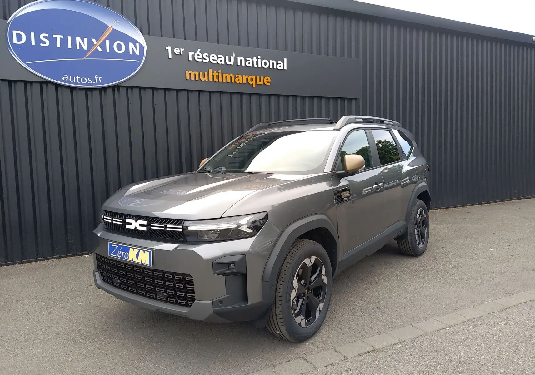 SUV Dacia Bigster gris schiste en 3/4 avant droit, avec jantes noires et rétroviseurs beige devant un mur noir.