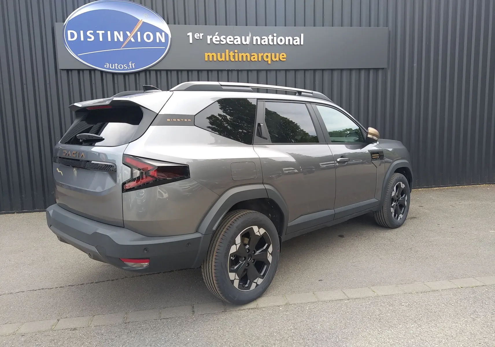 Vue 3/4 arrière droite du Dacia Bigster gris schiste avec jantes noires et détails bronze, stationné devant un mur sombre.