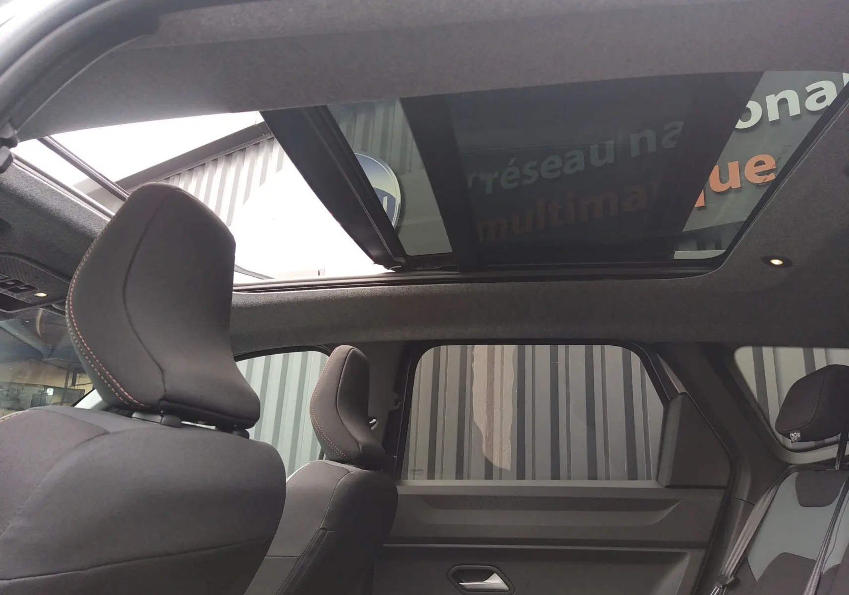 Vue intérieure arrière du Dacia Bigster gris schiste 2025, mettant en valeur le toit panoramique et les sièges noirs avec surpiqûres rouges.