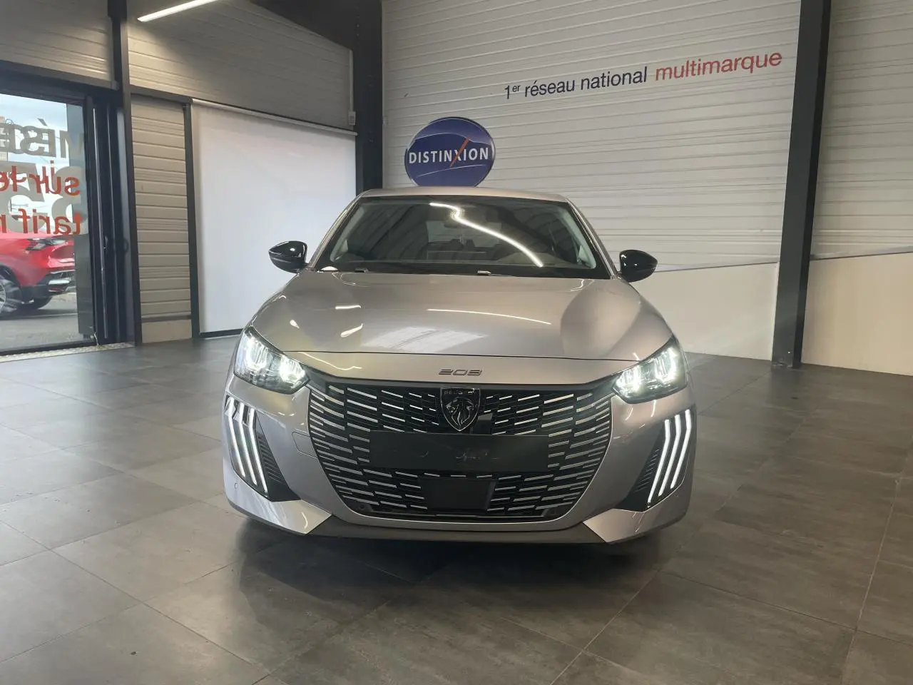 Vue frontale d'une Peugeot 208 1.2i Hybrid gris Artense avec phares LED et calandre noire brillante en showroom.