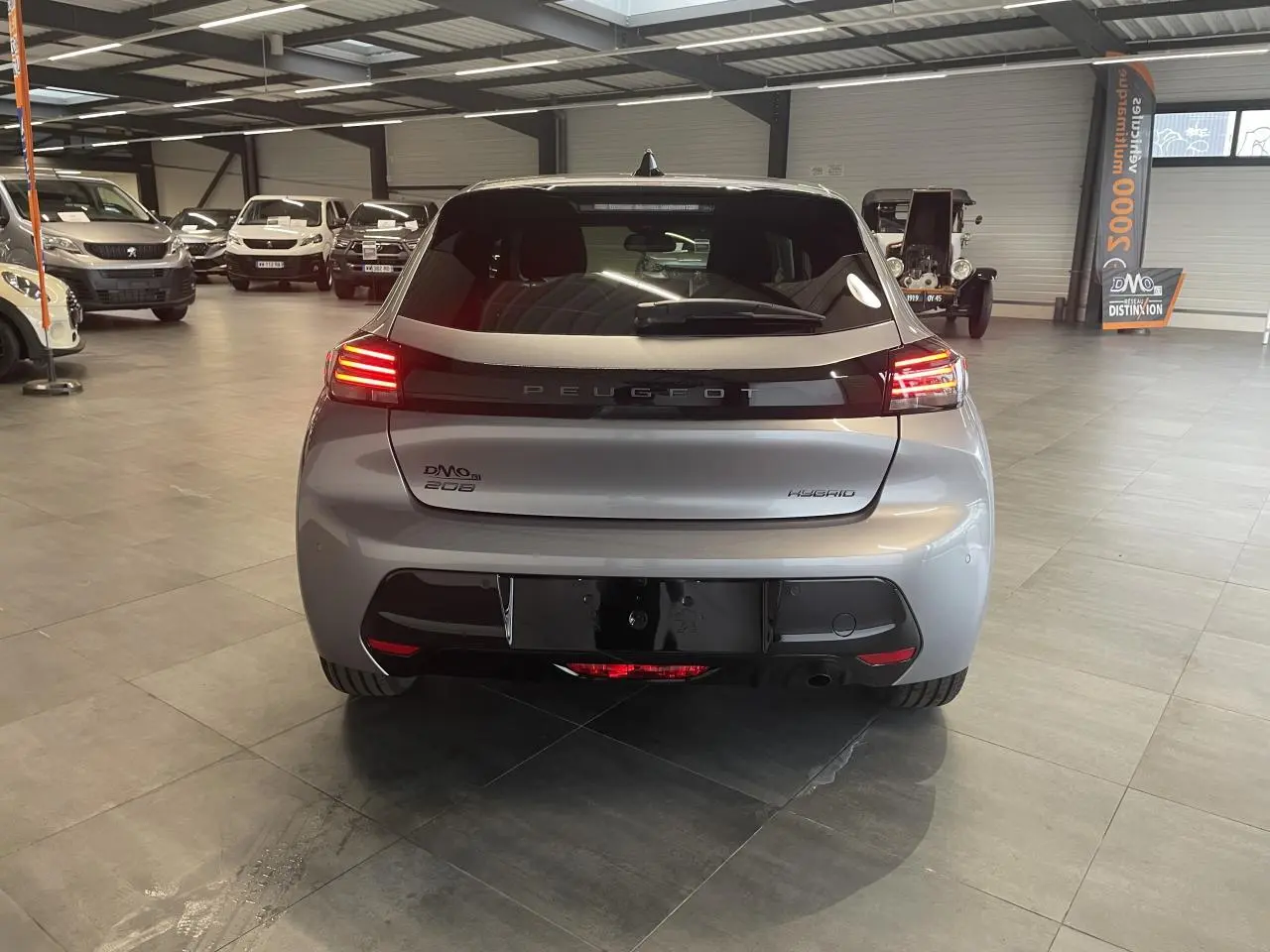 Vue arrière d'une Peugeot 208 hybride gris Artense avec feux arrière LED allumés en intérieur showroom.