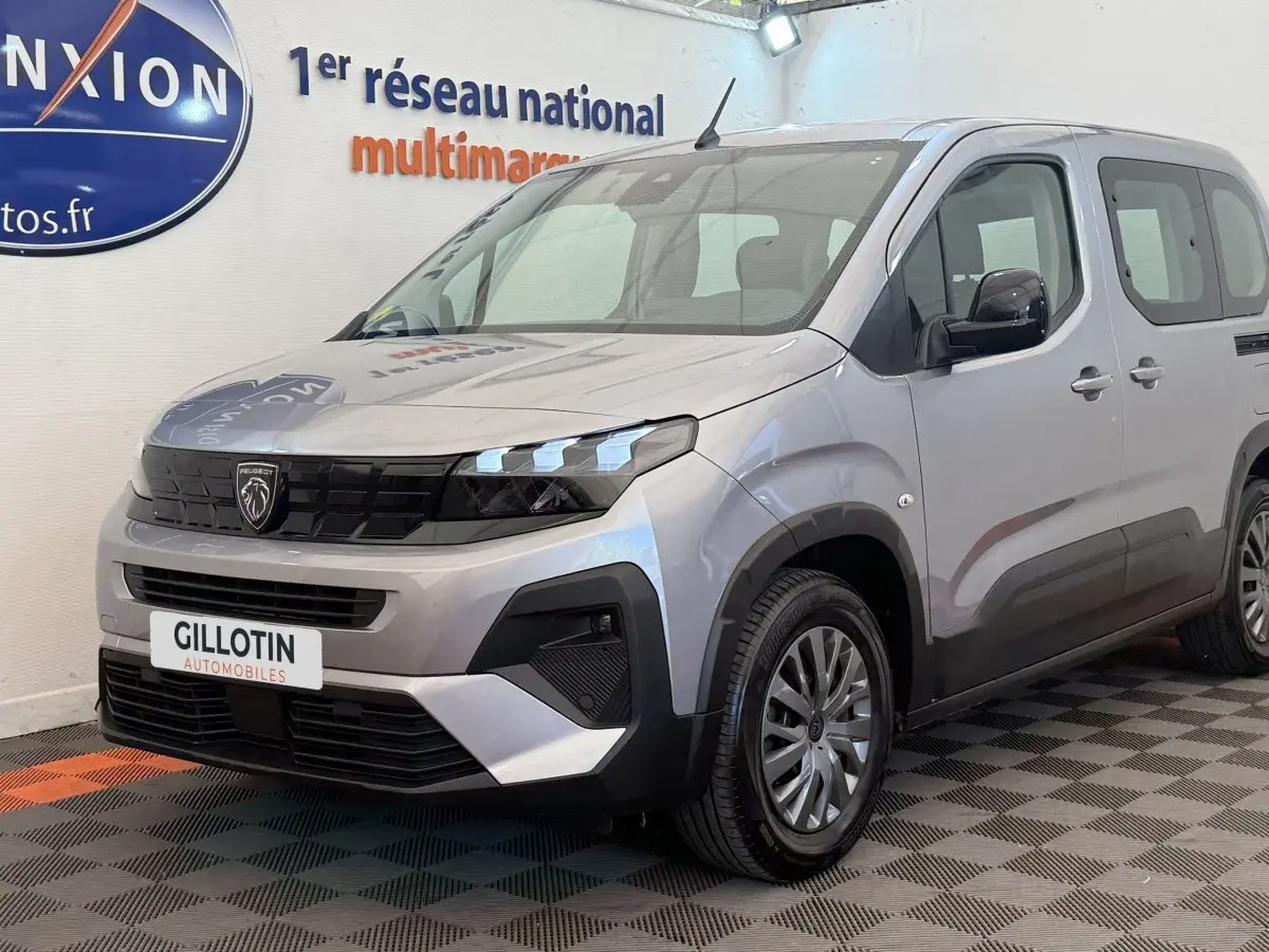 Peugeot Rifter gris clair en 3/4 avant droit, avec calandre noire et feux LED distinctifs, stationné en intérieur.