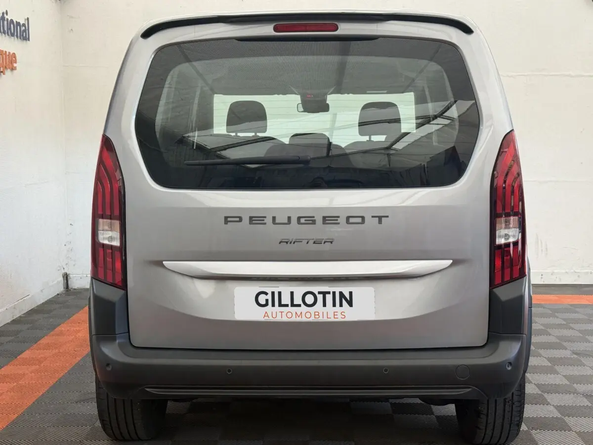 Vue arrière du Peugeot Rifter gris clair 2024 avec feux verticaux et pare-chocs noir mat visible en intérieur.