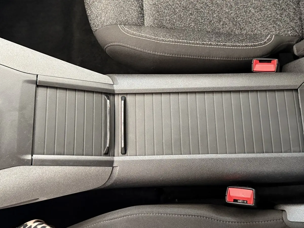 Gros plan sur la console centrale grise du Peugeot Rifter 2024, avec sièges tissu et boucles de ceinture rouges visibles.