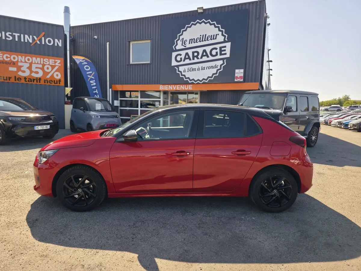 Opel Corsa rouge vue de profil côté gauche, avec toit noir et jantes alliage noires, stationnée devant un garage.