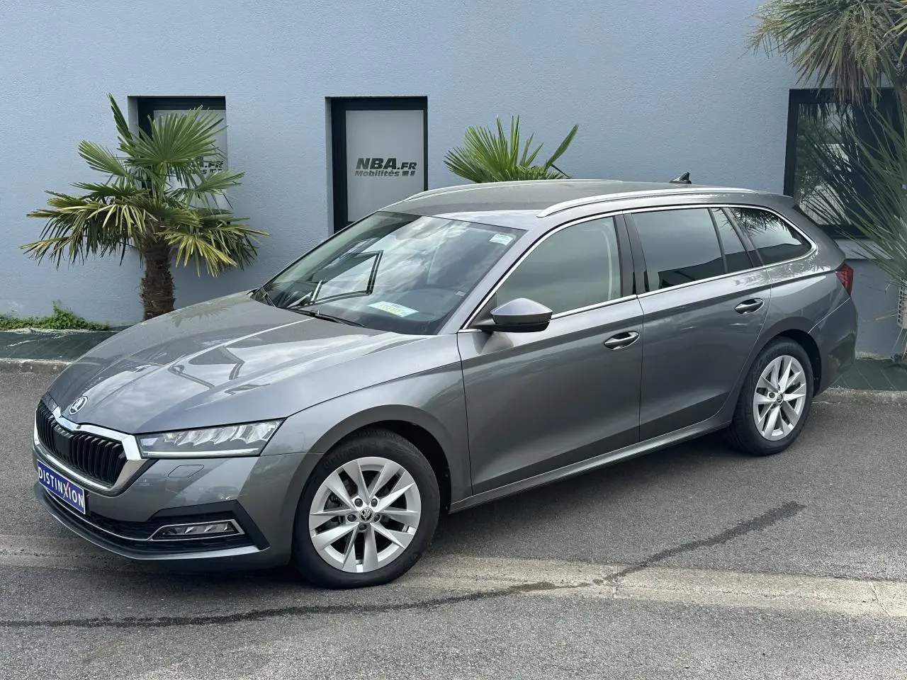 Skoda Octavia Combi gris graphite vue 3/4 avant droit, avec jantes alliage et vitres teintées, stationnée devant un mur bleu.
