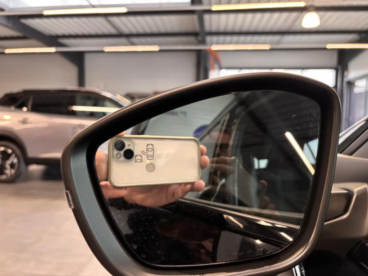 Rétroviseur côté droit du Peugeot 2008 noir, avec reflet partiel de l'habitacle et main tenant un smartphone.