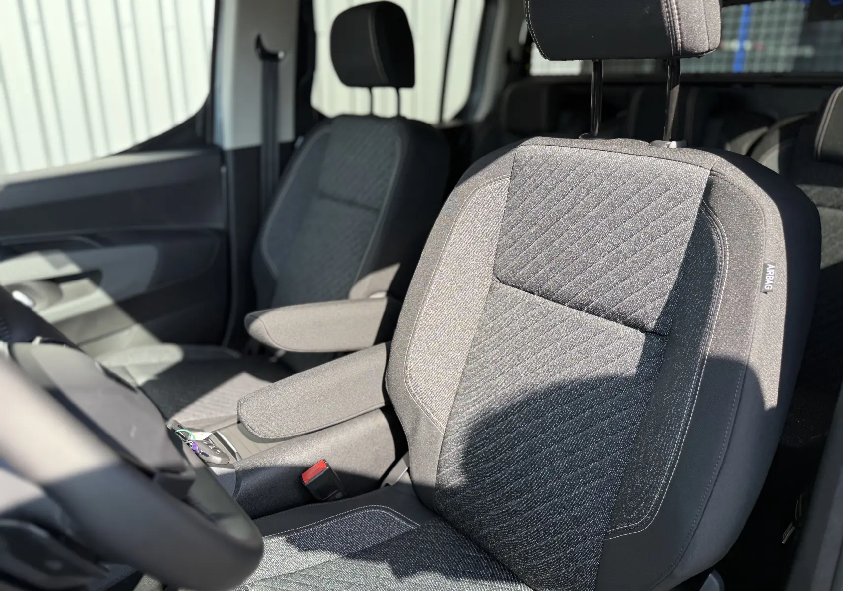 Gros plan sur les sièges avant gris foncé du Peugeot Rifter Allure 2026, avec accoudoirs et étiquette airbag visible.