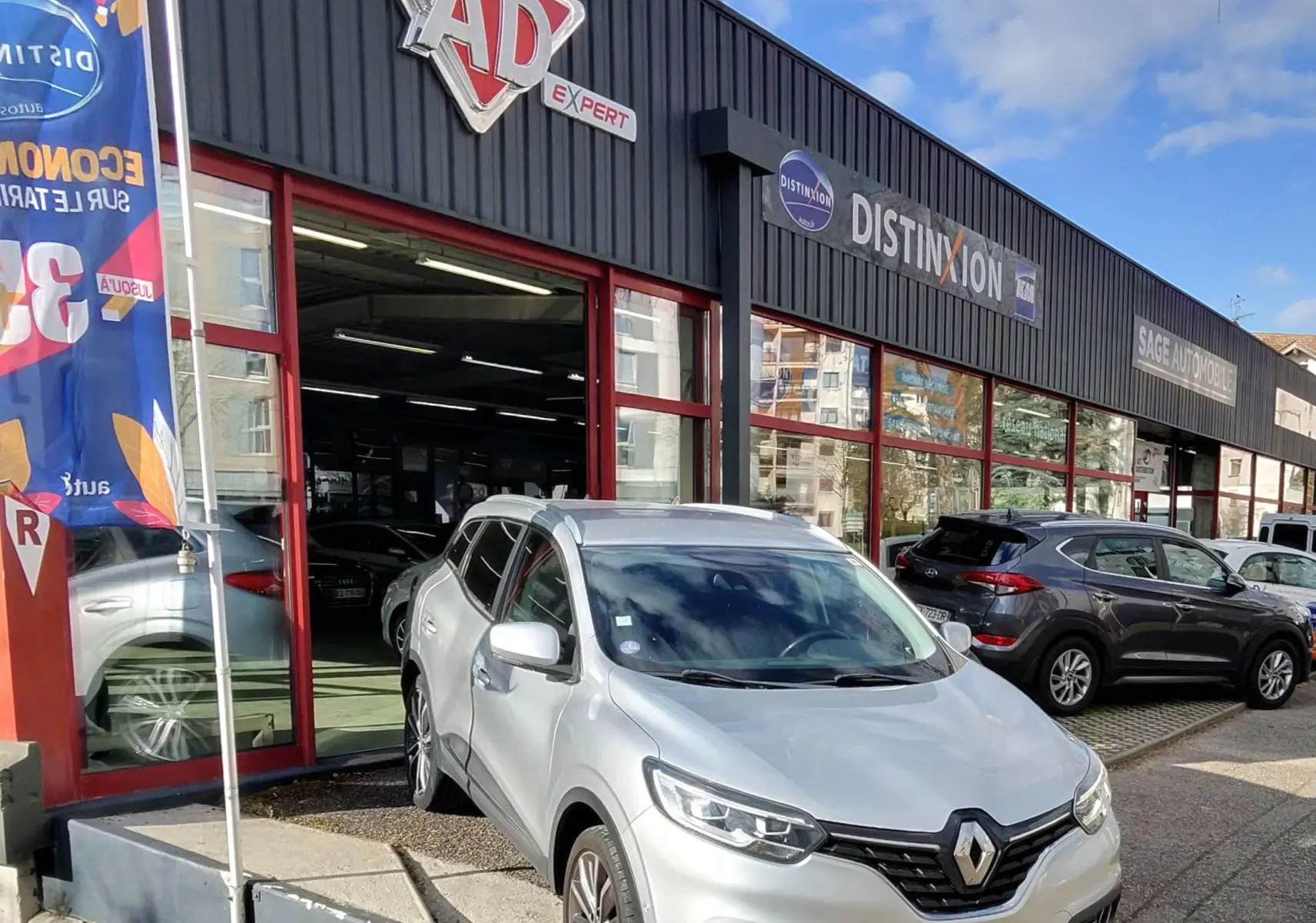 Renault Kadjar gris platine métal en 3/4 avant, stationné devant un garage avec enseignes visibles.