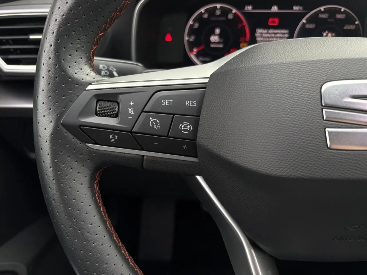 Gros plan sur le volant cuir noir perforé avec surpiqûres rouges du SEAT LEON 2024, boutons de régulateur de vitesse visibles.