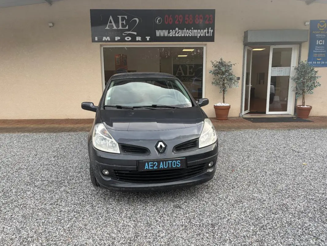 Vue avant d'une Renault Clio III gris foncé, avec plaque AE2 AUTOS, stationnée devant un bâtiment commercial.