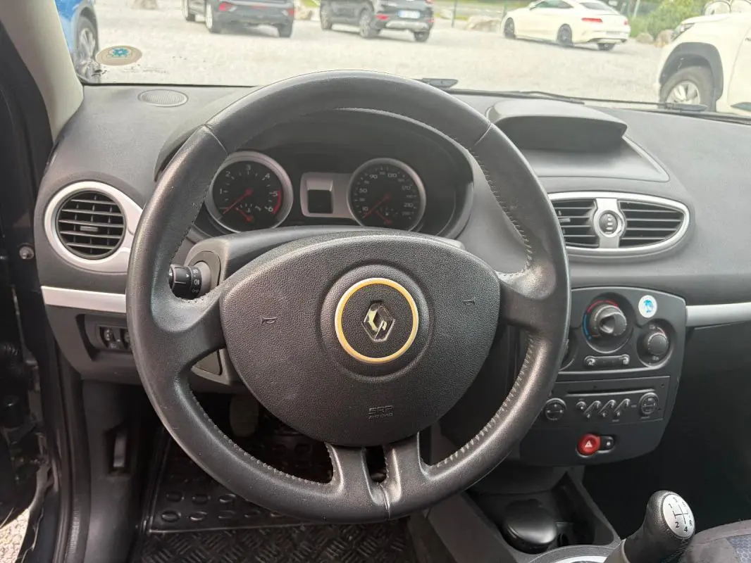 Vue intérieure centrée sur le volant noir de la Renault Clio III gris, avec tableau de bord et levier de vitesse manuel visibles.