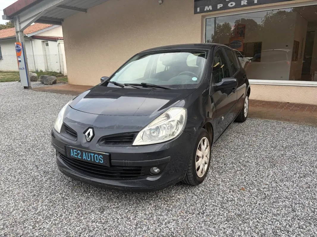 Renault Clio III gris vue 3/4 avant droit, avec plaque AE2 AUTOS, garée sur gravier devant un bâtiment.