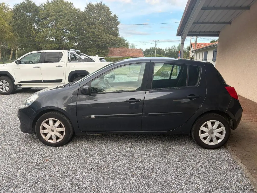 Renault Clio III gris vue de profil côté gauche, garée sur gravier devant un abri et un mur beige.