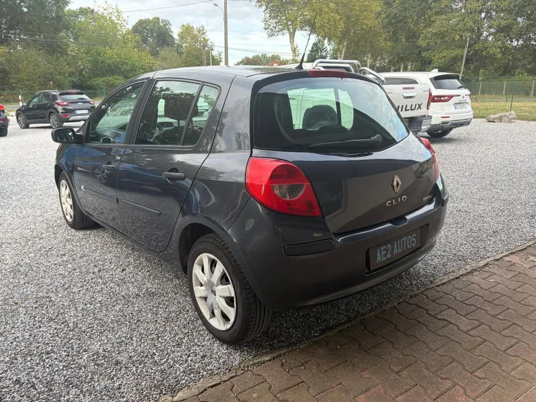 Renault Clio III gris vue 3/4 arrière droit, avec feux arrière rouges et plaque AE2 AUTOS visible.