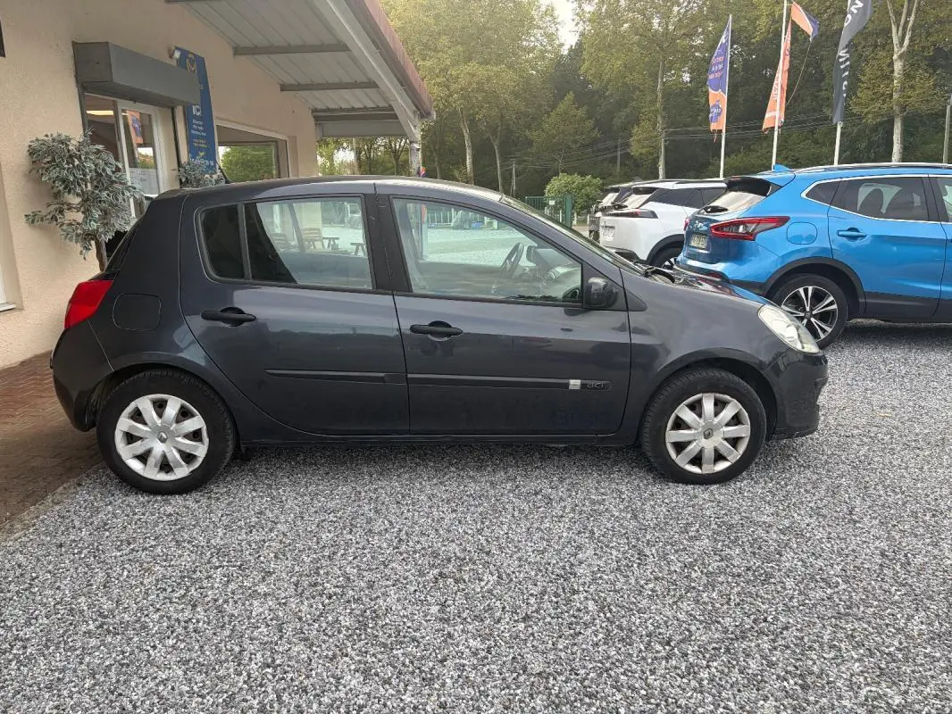 Profil côté droit d'une Renault Clio III gris foncé de 2005, garée sur un sol gravillonné devant un bâtiment.