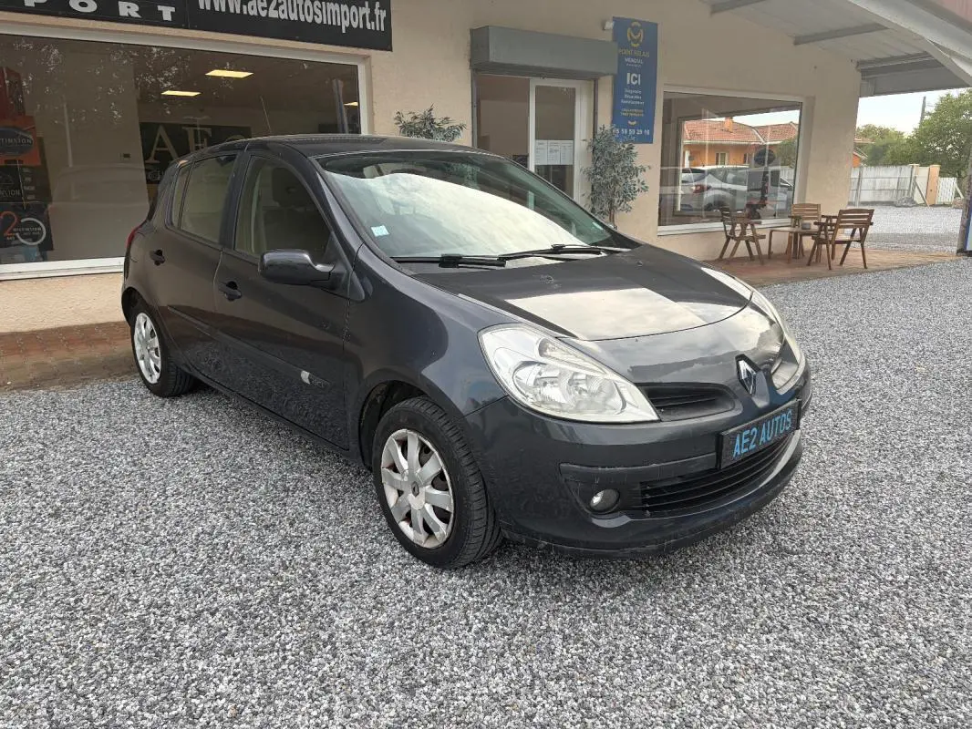Renault Clio III gris vue 3/4 avant droit, garée sur gravier devant un bâtiment avec vitrine et terrasse.