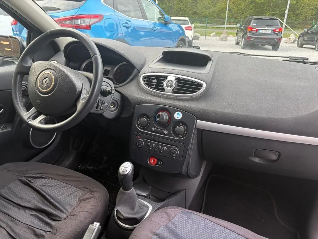 Vue intérieure côté conducteur de la Renault Clio III gris 2005, avec tableau de bord et levier de vitesses manuel visibles.
