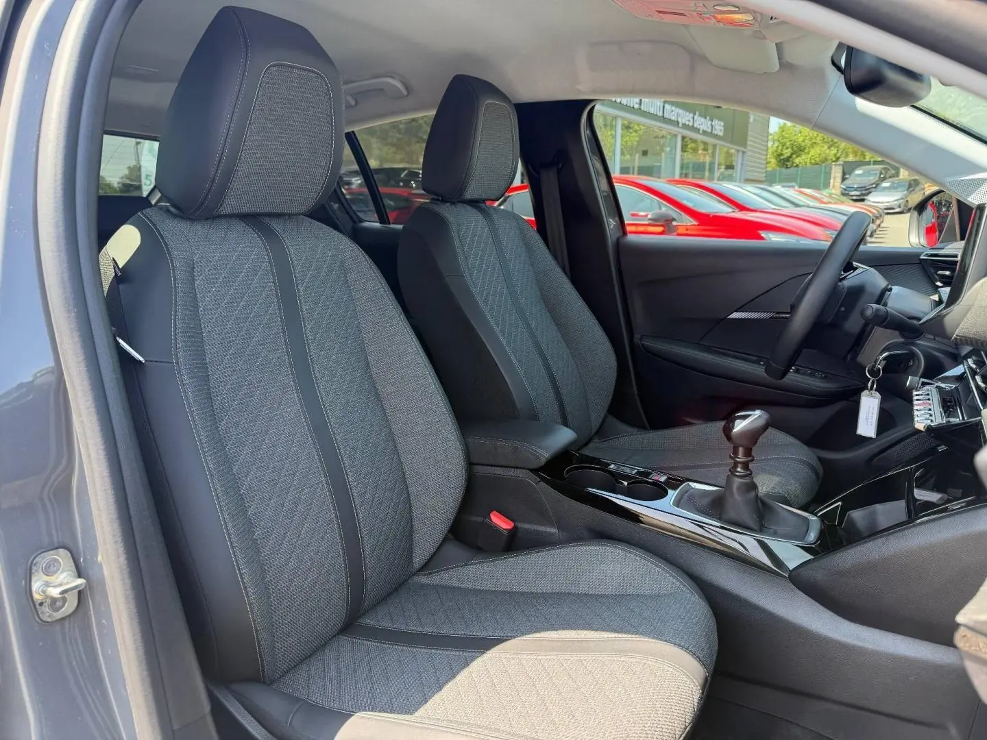 Vue intérieure côté conducteur de la Peugeot 208 gris Artense, sièges tissu gris et levier de vitesse manuel visible.