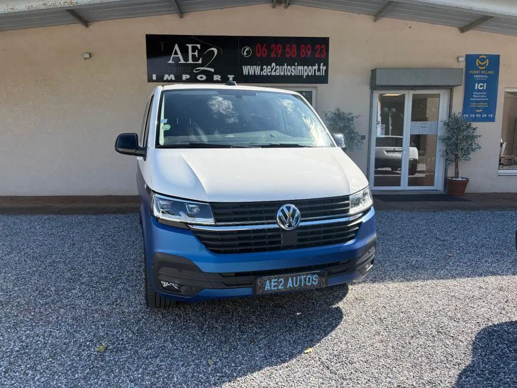 Vue avant d'un Volkswagen Transporter blanc avec pare-chocs bleu, stationné devant un bâtiment commercial.