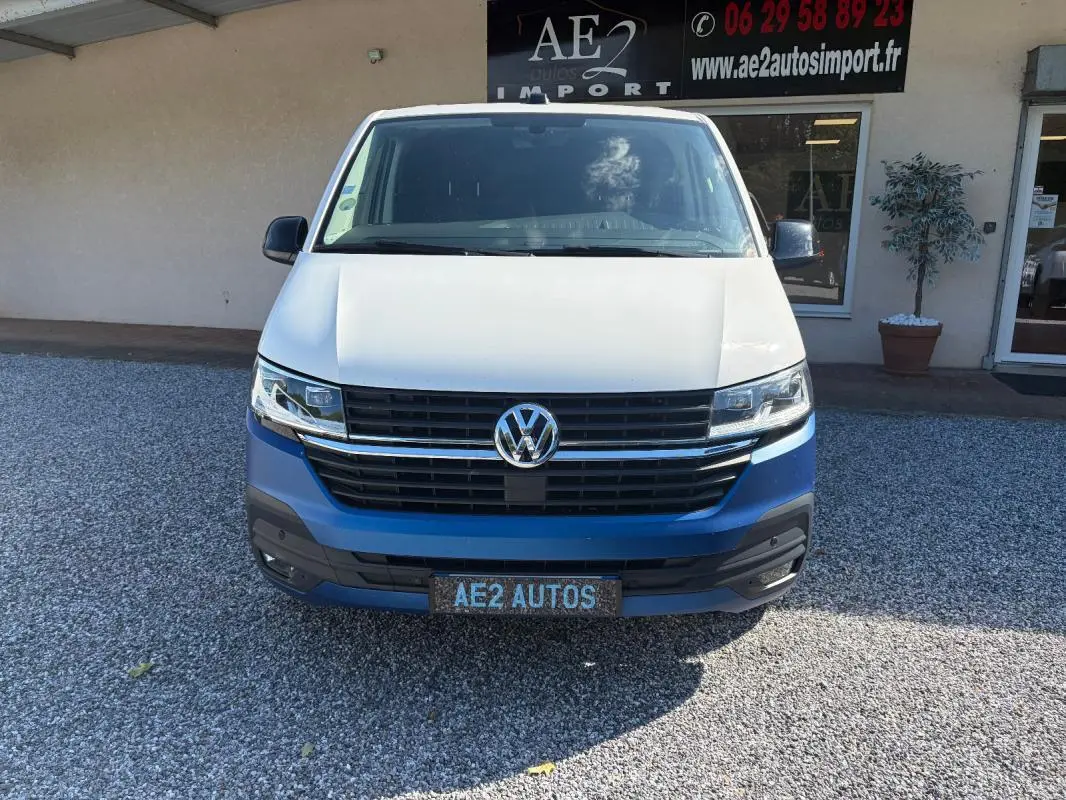 Vue avant du Volkswagen Transporter blanc avec calandre noire et logo VW bien visible, stationné devant un bâtiment.
