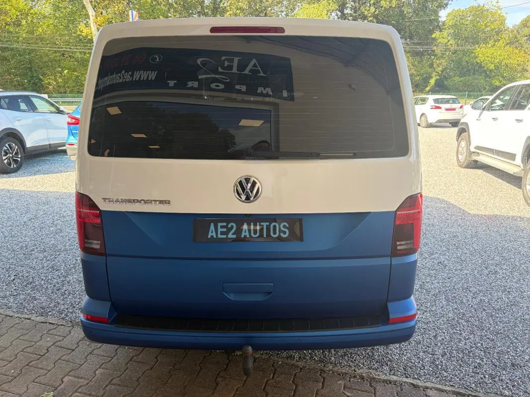 Vue arrière d’un Volkswagen Transporter blanc et bleu, avec vitre arrière teintée et plaque AE2 AUTOS visible.