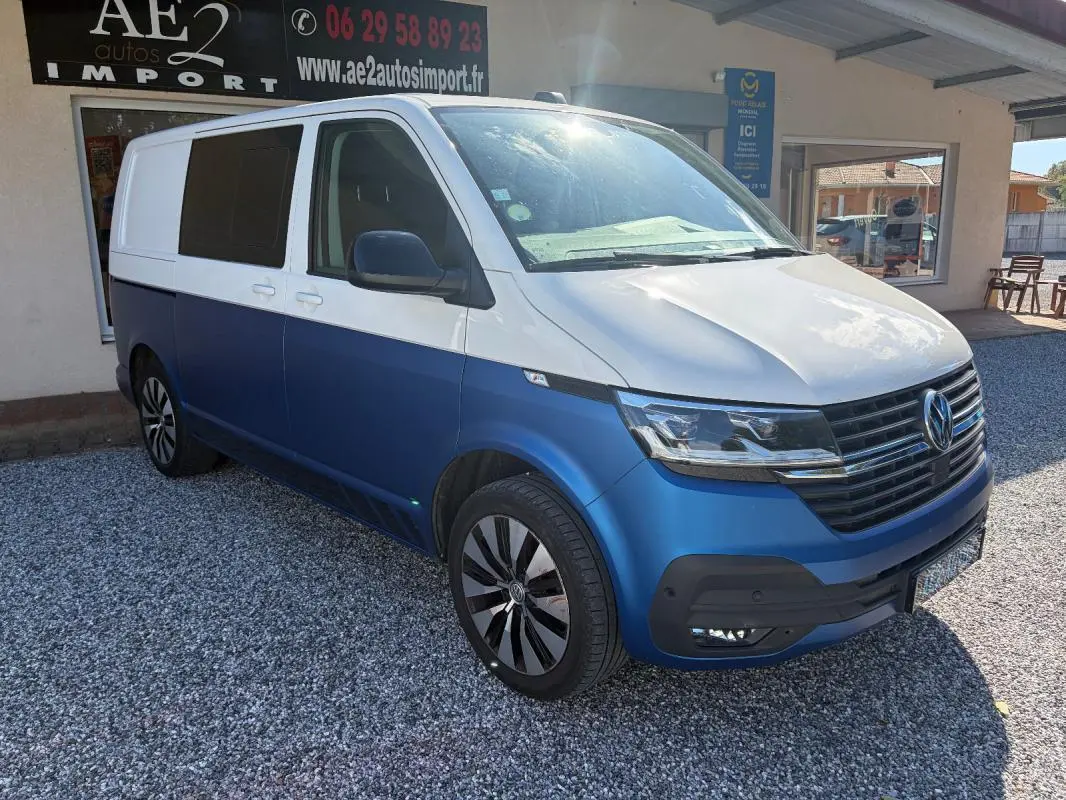 Vue 3/4 avant droit d’un Volkswagen Transporter blanc et bleu avec jantes aluminium, stationné devant un bâtiment.
