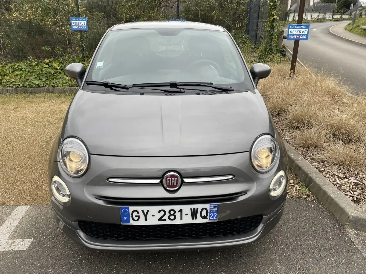 Vue avant d'une FIAT 500 gris foncé avec phares allumés, stationnée sur un parking réservé Rent A Car.