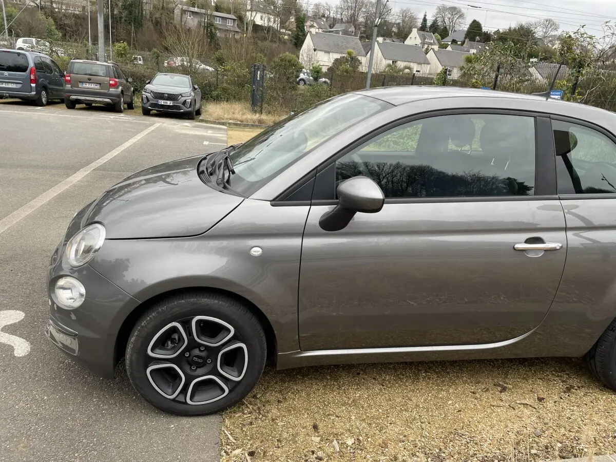 Vue de profil côté gauche d'une FIAT 500 Hybrid gris foncé avec jantes noires et détails chromés sur la poignée.