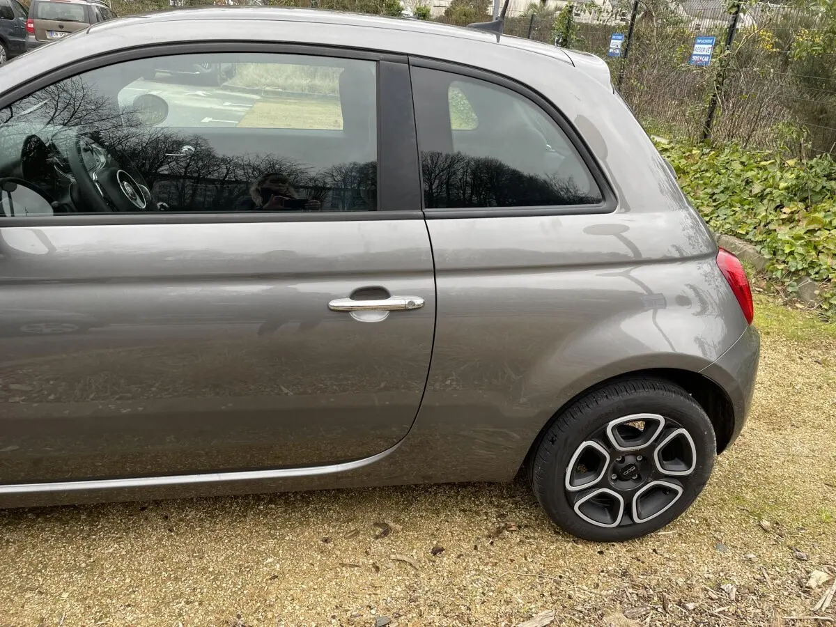 Vue de profil côté gauche d'une FIAT 500 gris foncé avec jante alliage distinctive, stationnée sur sol gravillonné.
